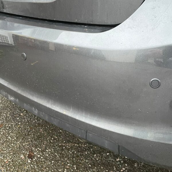 Close-up of a gray car's rear bumper with parking sensors and a partly visible license plate.
