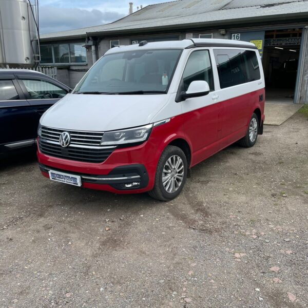 A red and white Volkswagen van parked on gravel outside an industrial building.