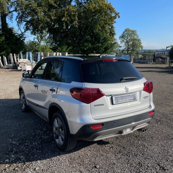 Rear view of a silver Suzuki Vitara parked on gravel with trees and an industrial area in the background.