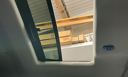 Partially open vehicle sunroof showing a ceiling with wooden beams and boxes above, and a control panel with blue buttons to the right.